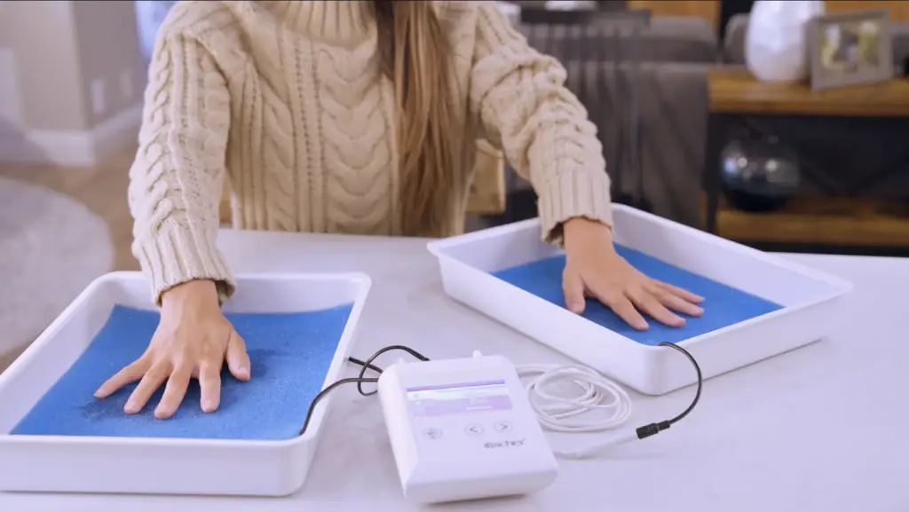 An overhead photograph of only the arms and hands of a person facing forward treating their palmar hyperhidrosis (excessive sweating of the hands) using RA Fischer Co.'s The Fischer iontophoresis device on a white table. White water bath trays and black, metal-free silicone electrodes with ph-balancing foam inside the tray on a plain white background. The Fischer iontophoresis device is white and rectangular with a blue and grey screen. Buttons read "SET" and arrows. Logo of the Fischer device bottom right corner