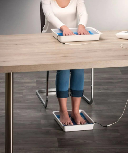 A woman dressed in a white blouse and jeans submerges her hands and feet into the Fischer iontophoresis device's water bath trays, aiming to alleviate palmar hyperhidrosis (excessive hand sweating) and plantar hyperhidrosis (excessive foot sweating). RA Fischer Co.'s Fischer iontophoresis device enables the simultaneous treatment of both hands and feet.