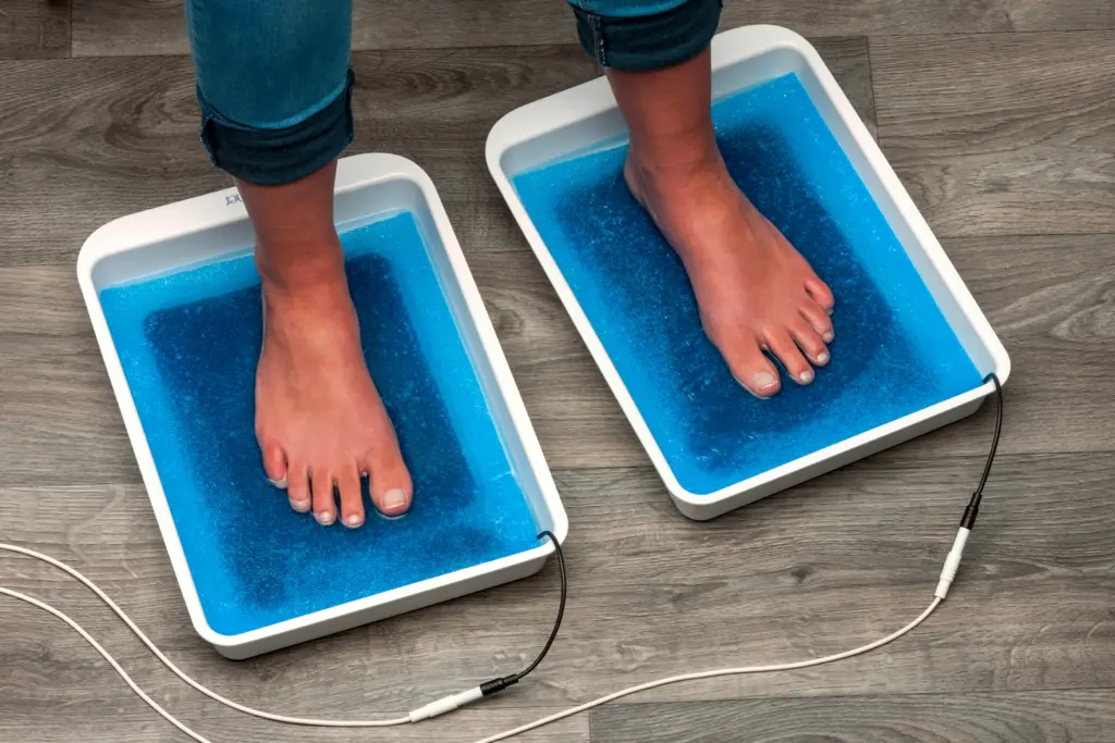 An aerial view captures the therapeutic journey of an individual who is diligently addressing the challenges of plantar hyperhidrosis, characterized by excessive sweating of the feet, with the assistance of RA Fischer Co.'s innovative solution, The Fischer iontophoresis device. Positioned on a warm and inviting wooden floor, the individual's feet rest comfortably in separate white trays, thoughtfully designed to contain blue foam within. This meticulous setup signifies a conscious effort to provide a holistic and effective approach to managing this condition, demonstrating the commitment of RA Fischer Co. to improving the lives of those affected by hyperhidrosis.